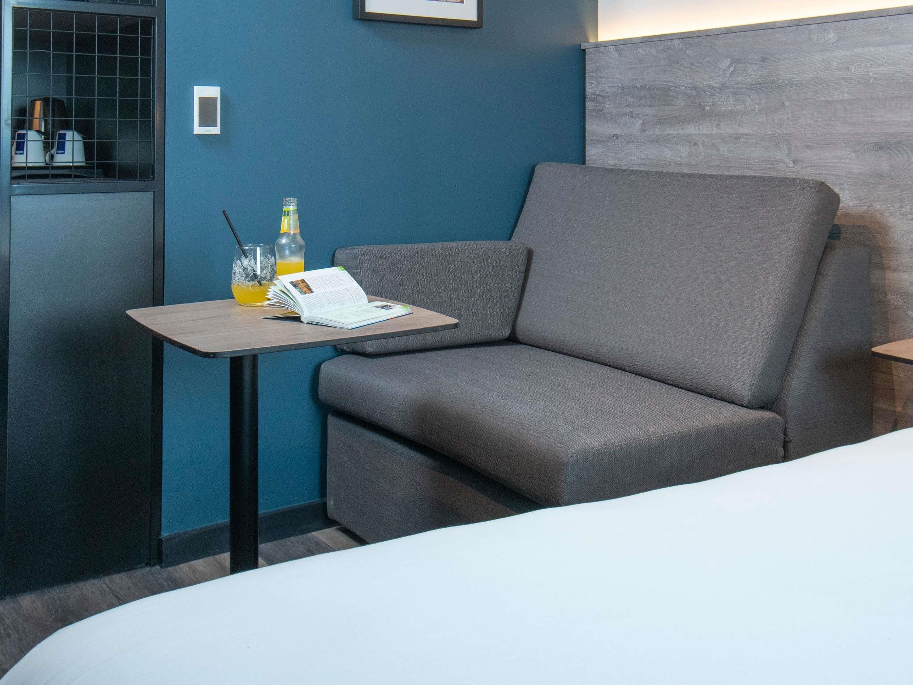 A modern hotel room with a grey sofa, a small table with a book and a bottle, and a minibar.