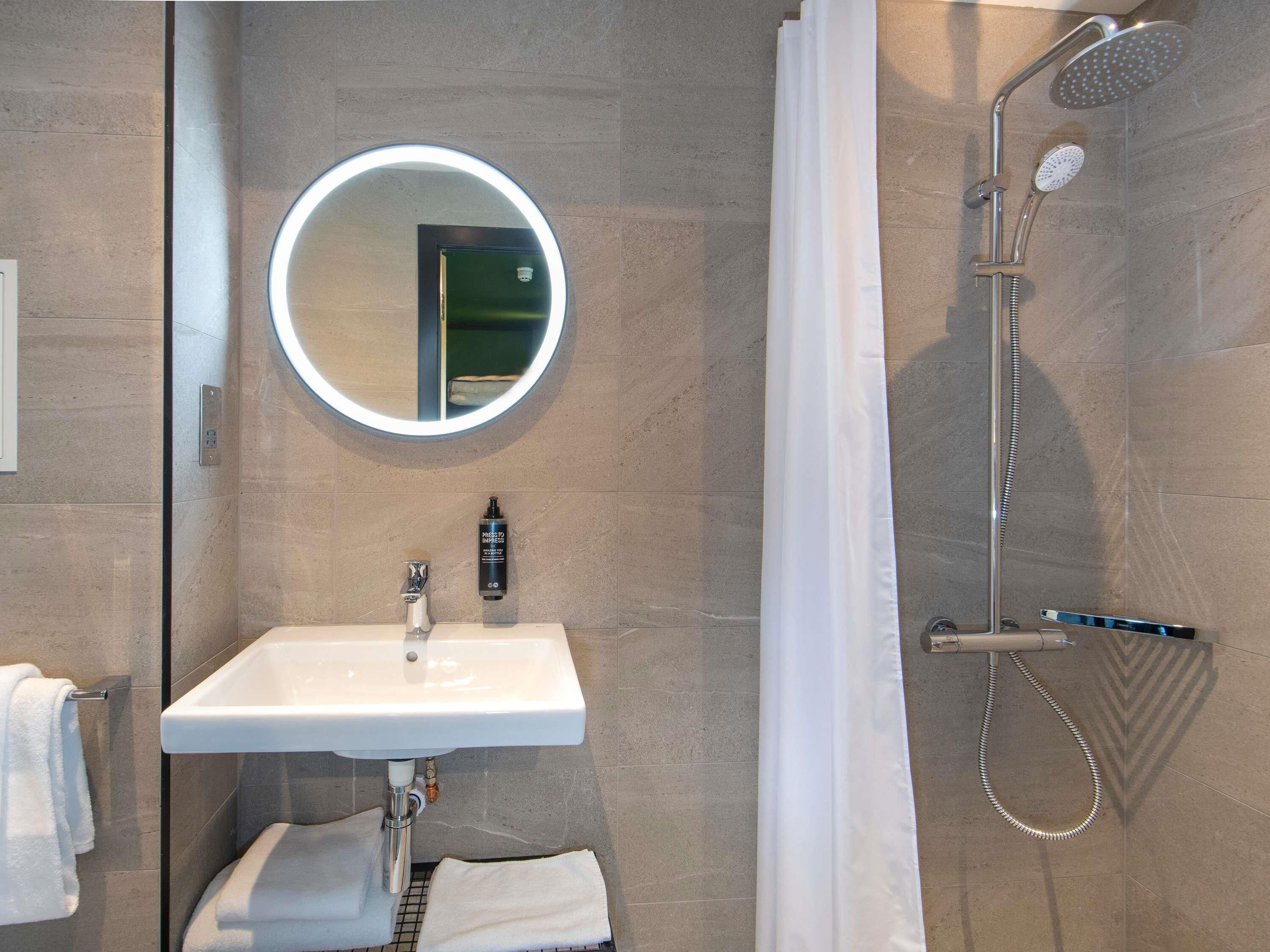 A modern bathroom with a round mirror, a sink, a shower with a handheld showerhead, and a towel rack.