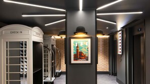 Un hall d’hôtel moderne avec des comptoirs d’enregistrement, une affiche encadrée représentant Londres et un écran numérique indiquant l’étage de l’ascenseur.