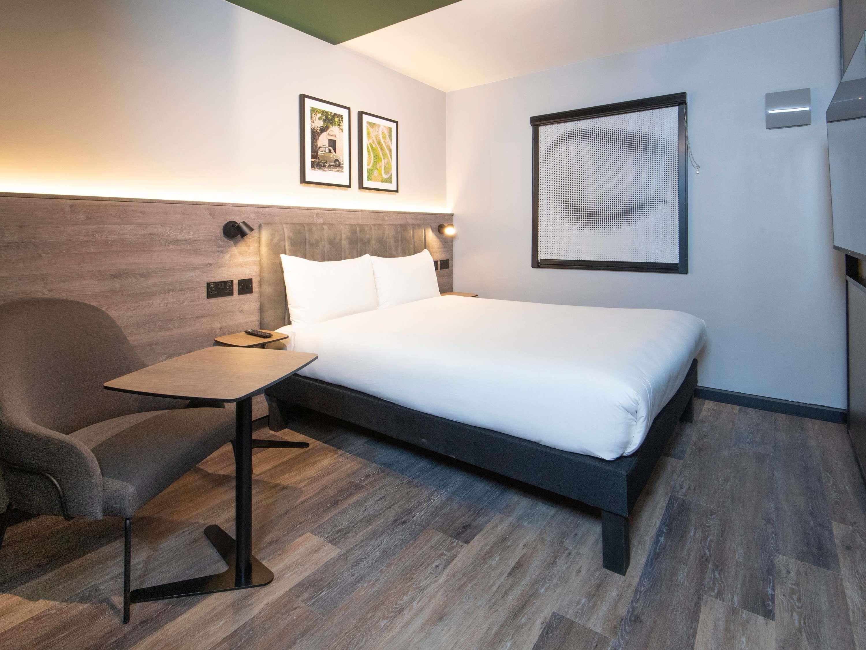 A hotel room with a bed, a chair, a small table, and two framed pictures on the wall.