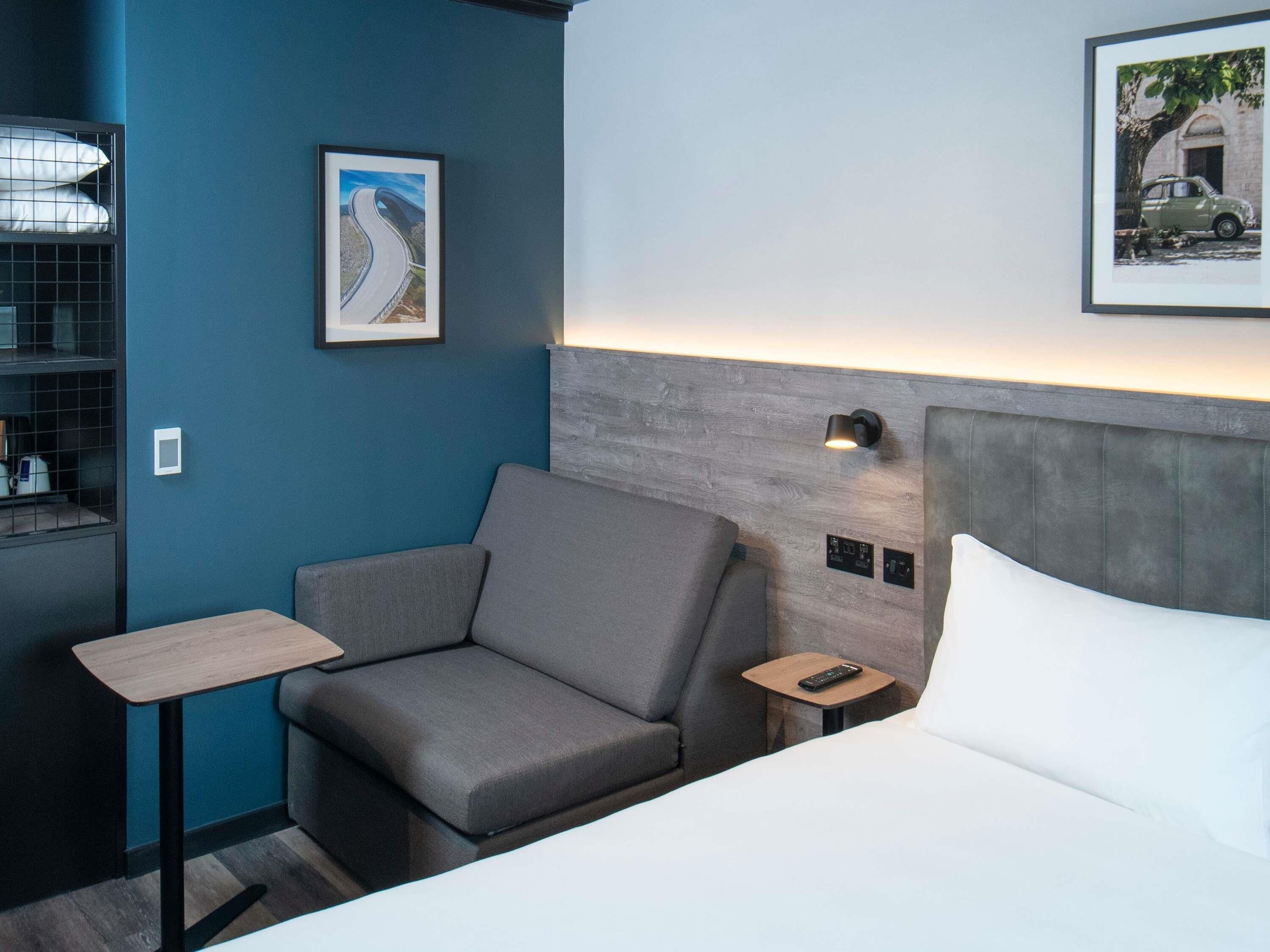 Hotellihuone, jossa on sänky, harmaa sohva, pieni pöytä, yöpöytä, seinään kiinnitetty lamppu ja kehystetty kuva mutkaisesta tiestä.