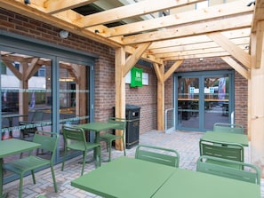 An outdoor seating area with green metal tables and chairs, a brick wall, and a wooden pergola structure. - Ibis Styles London Gatwick Airport (Crawley)