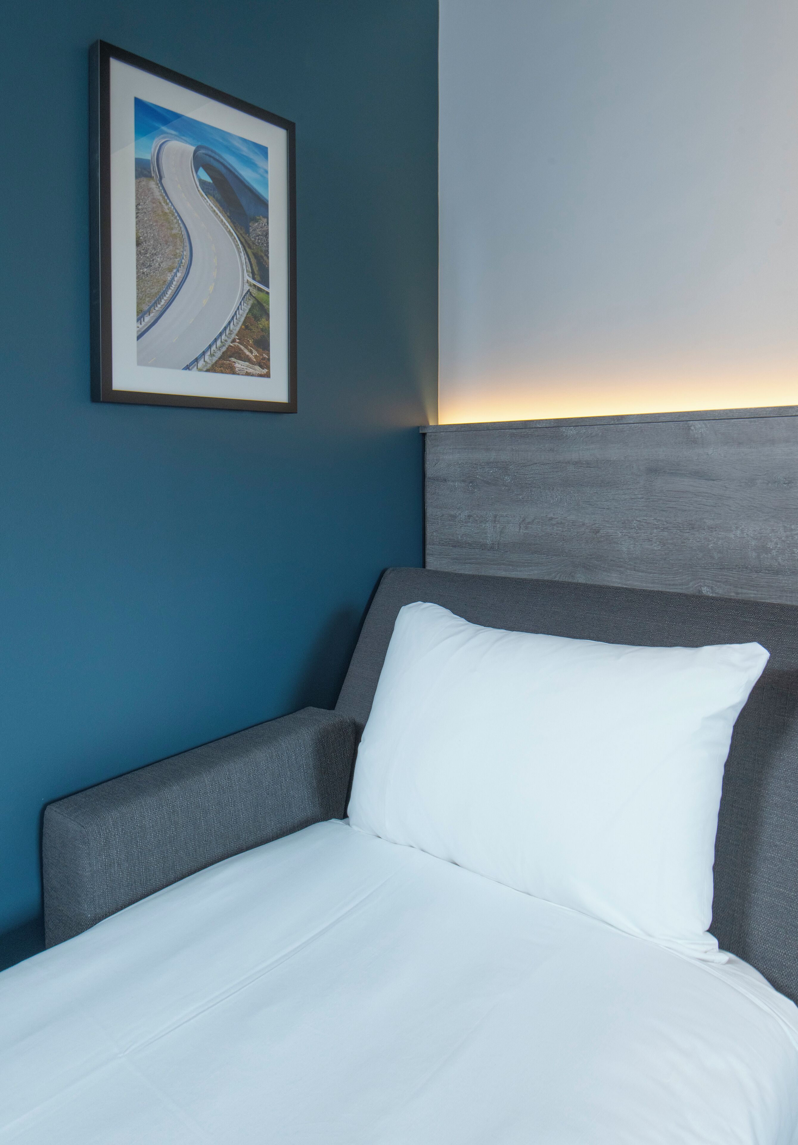A neatly made bed with white bedding, a grey headboard, and a framed picture of a winding road on the wall.