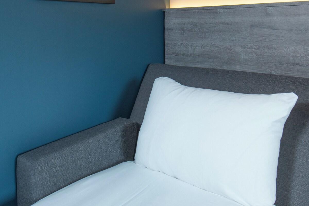 Una cama bien hecha con ropa de cama blanca, un cabecero gris y una foto enmarcada de un camino sinuoso en la pared.