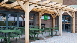 An outdoor seating area with green tables and chairs, a wooden pergola, and a sign for 'Ibis Styles'.