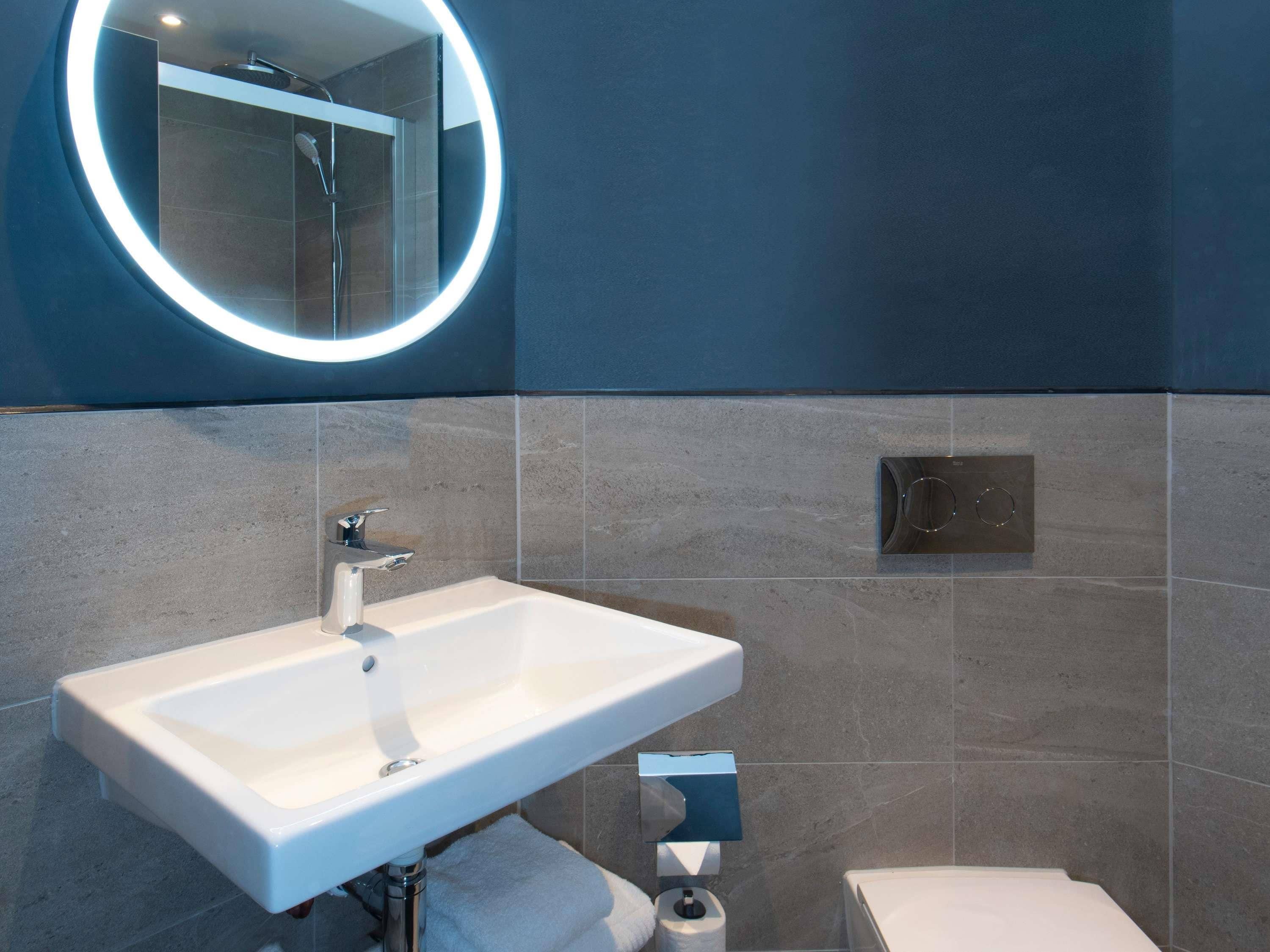Une salle de bain moderne avec un lavabo blanc, un miroir rond avec un éclairage intégré et un espace douche.