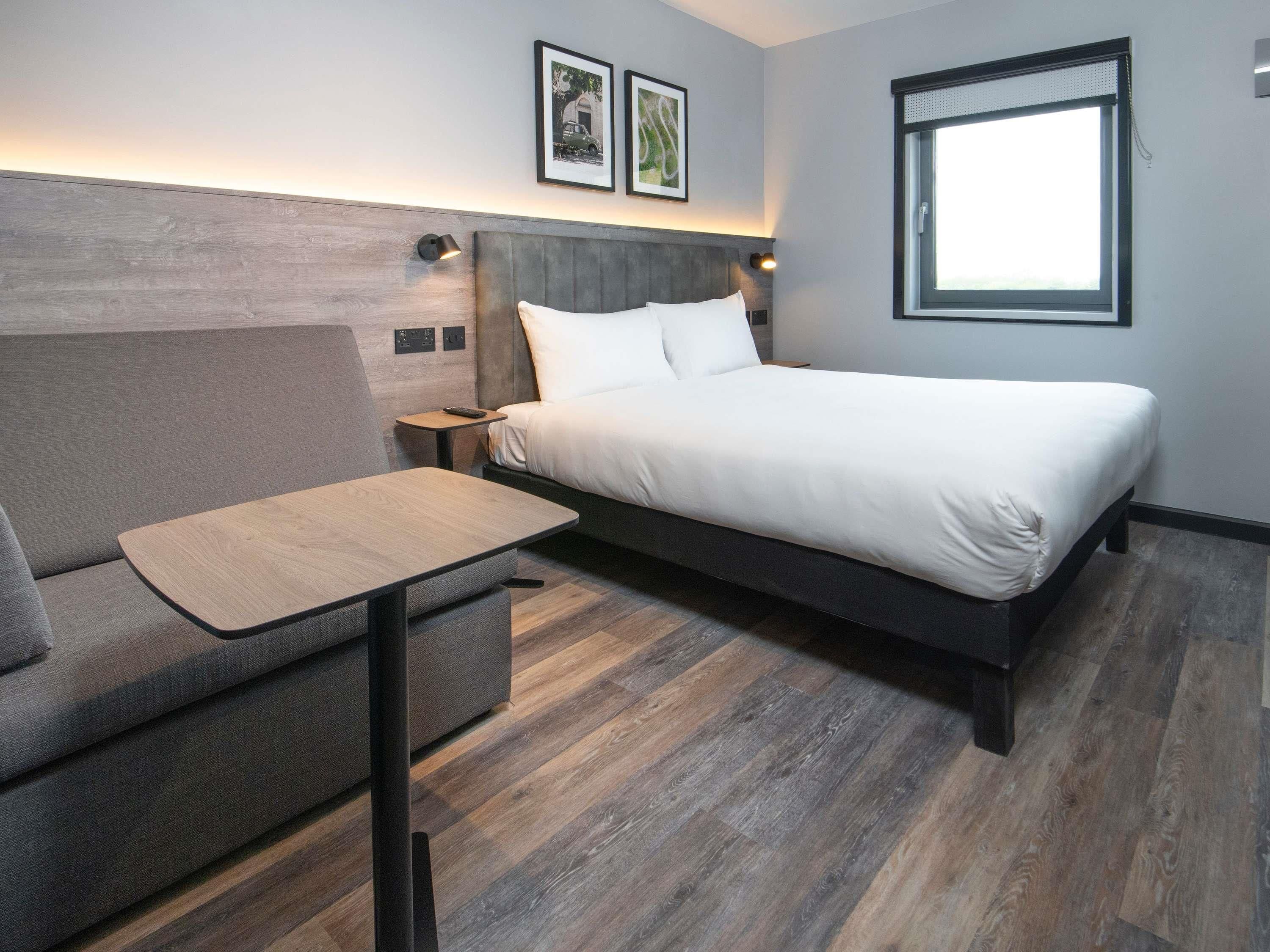 A modern hotel room with a bed, a sofa, a small table, and a window.