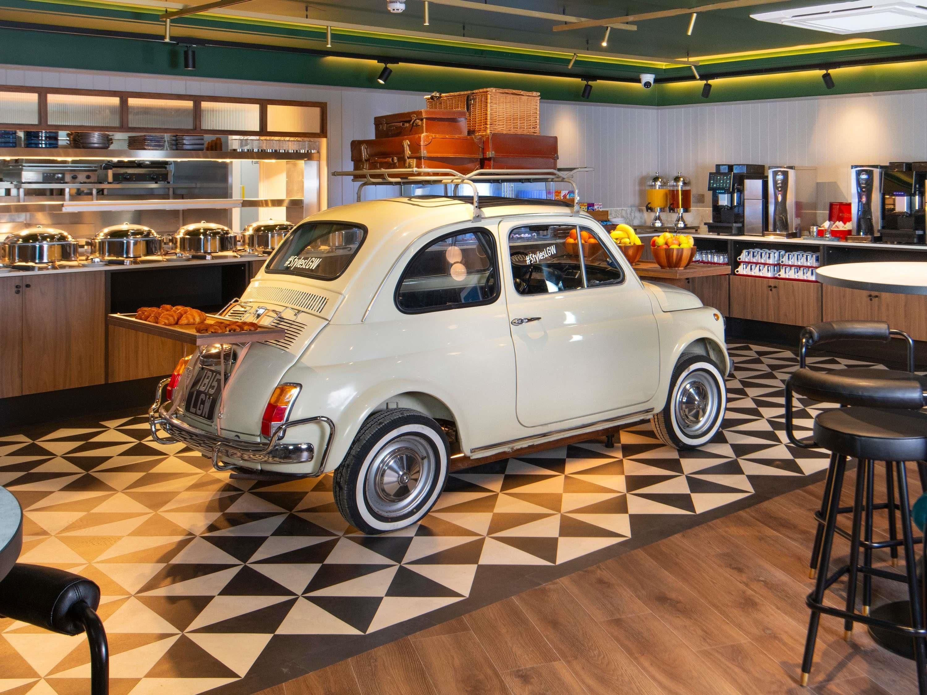 A vintage car parked in a modern kitchen with geometric patterned flooring and bar stools.
