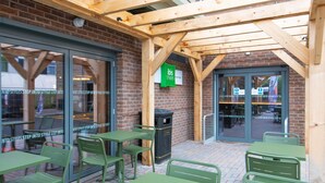 An outdoor seating area with green metal tables and chairs, a brick wall, and a wooden pergola structure.