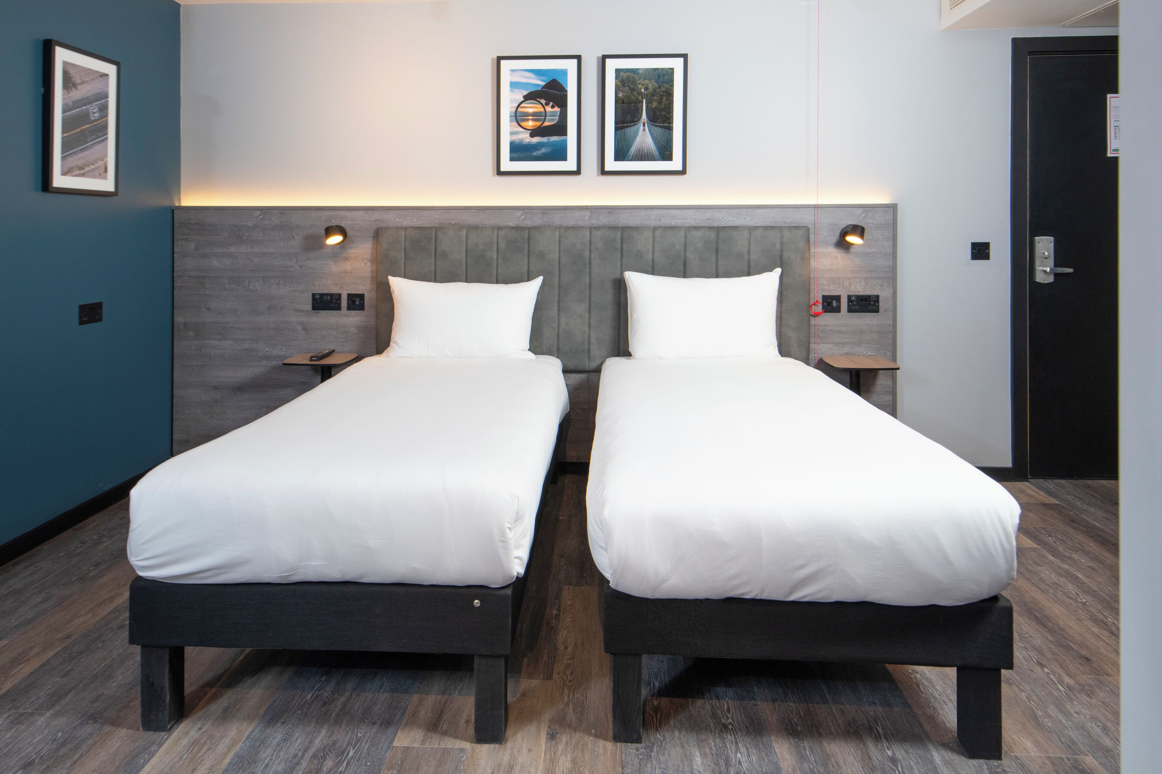 A hotel room with two single beds, a wooden headboard, bedside lamps, and framed pictures on the wall.