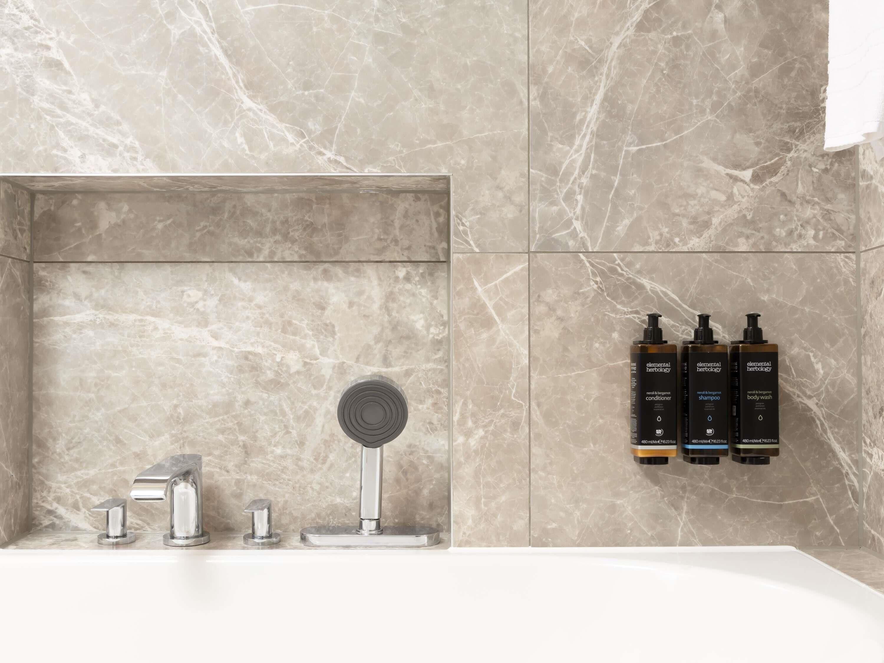 A bathroom with a marble wall, a shower with a modern faucet, and three bottles of Elemental Herbology body wash.