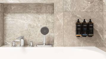 A bathroom with a marble wall, a shower with a modern faucet, and three bottles of Elemental Herbology body wash.