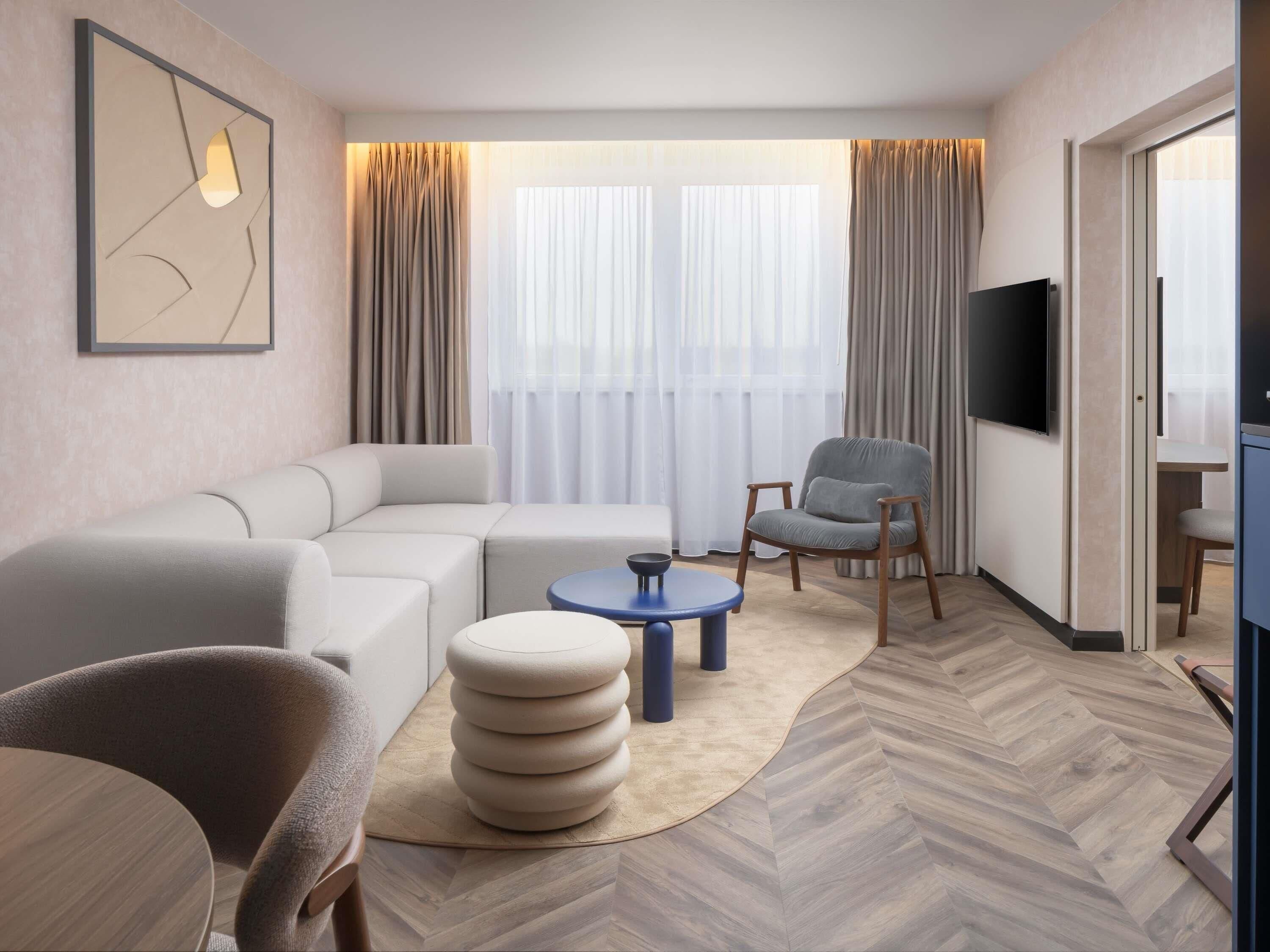 A modern hotel room with a sectional sofa, a round coffee table, a grey armchair, a flat-screen TV, and a herringbone-patterned floor.