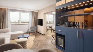 A modern hotel room with a sofa, chairs, a dining table, a kitchen area, and a TV.