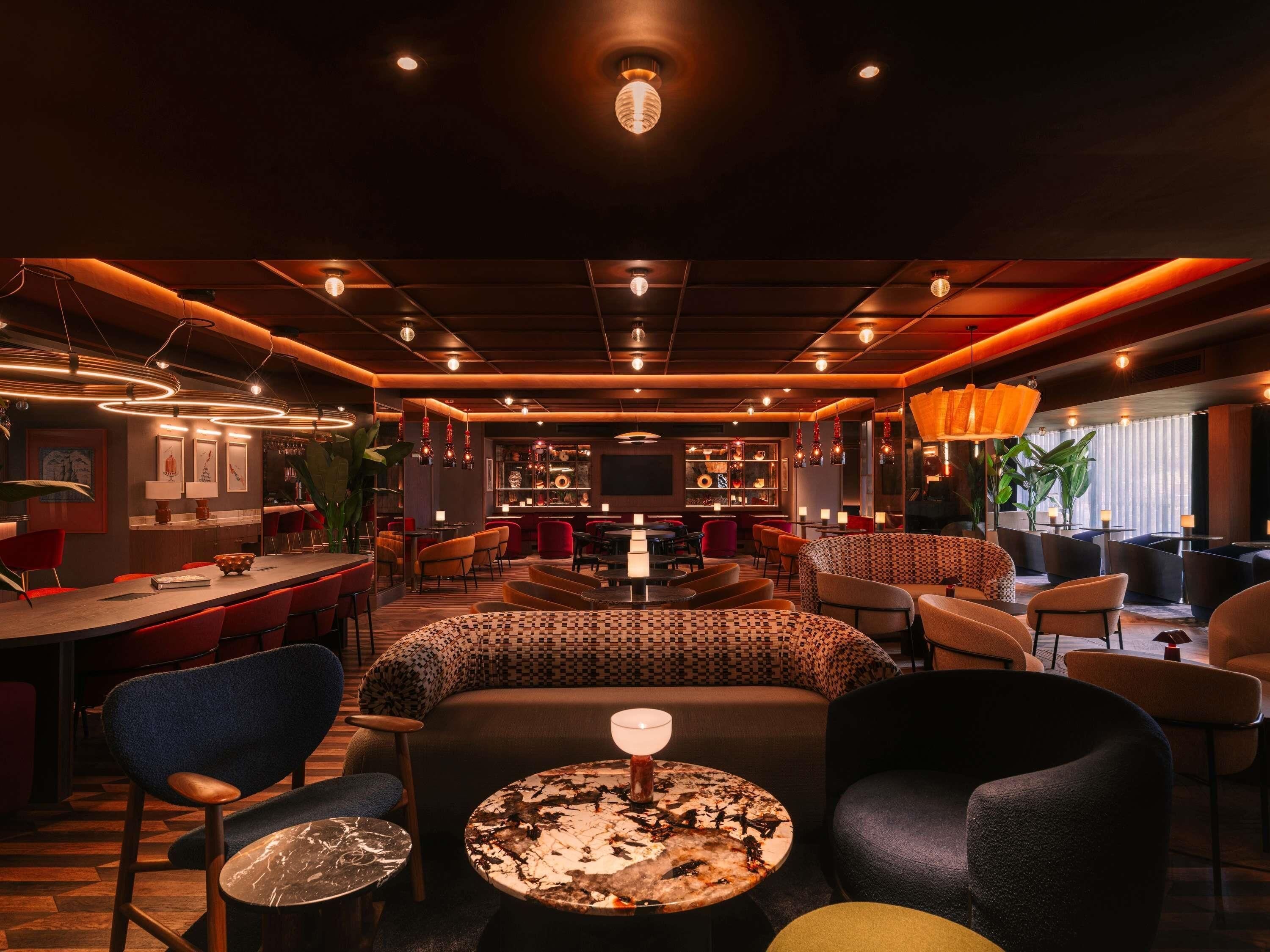 A modern bar area with a long counter, multiple seating options including bar stools and cushioned chairs, and a television screen mounted on the wall.