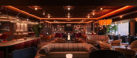 A modern bar area with a long counter, multiple seating options including bar stools and cushioned chairs, and a television screen mounted on the wall.