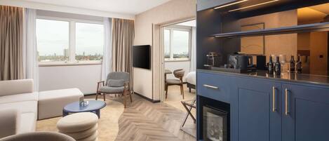 Una habitación de hotel moderna con sofá, sillones, una mesa de comedor, televisor, zona de cocina con cafetera y chimenea.