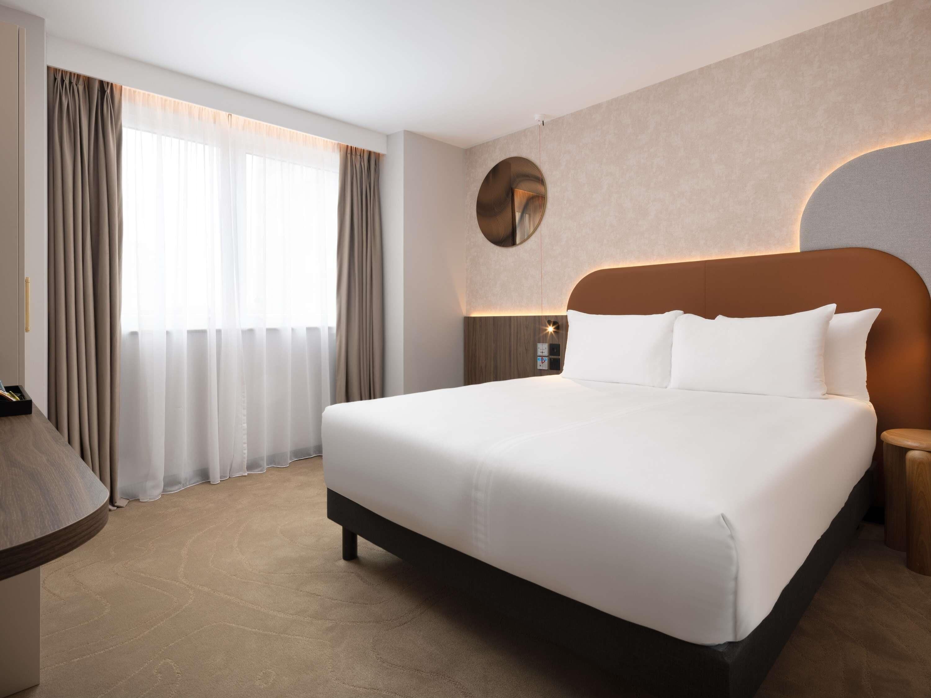A hotel room with a large bed, a bedside table, a lamp, and a view of the outdoors through the window.