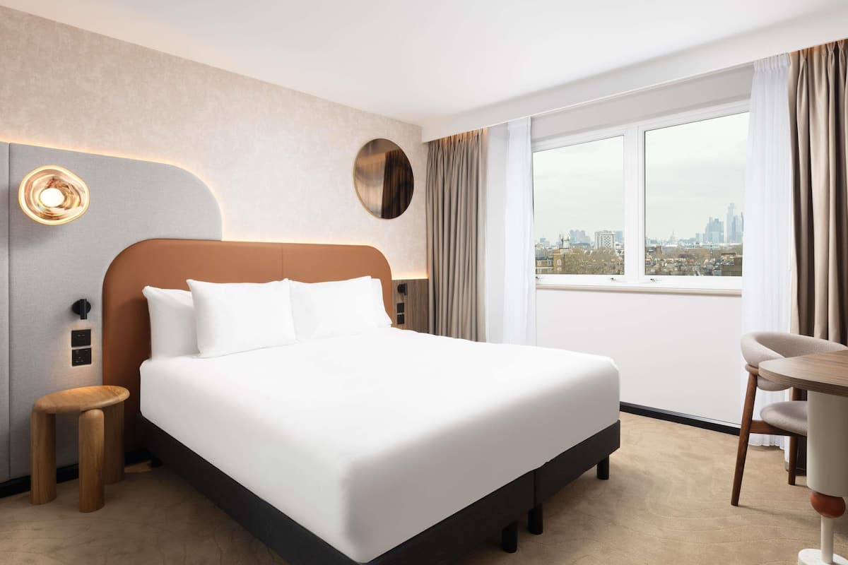 A modern hotel room with a large bed, a round wall clock, a city view, and a small table with a chair.
