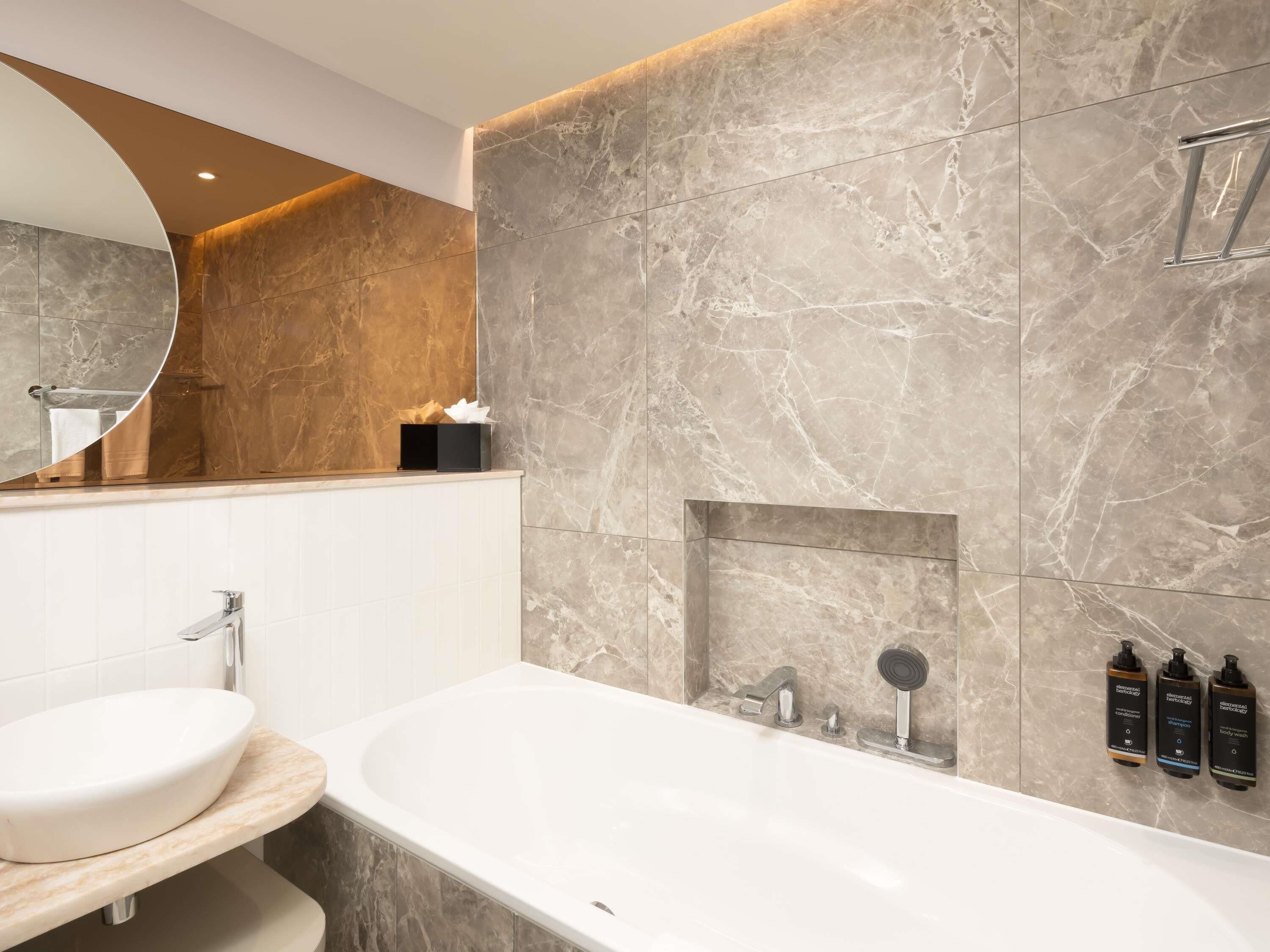 A modern bathroom with a marble wall, a freestanding bathtub, a round mirror, and a small shelf with toiletries.