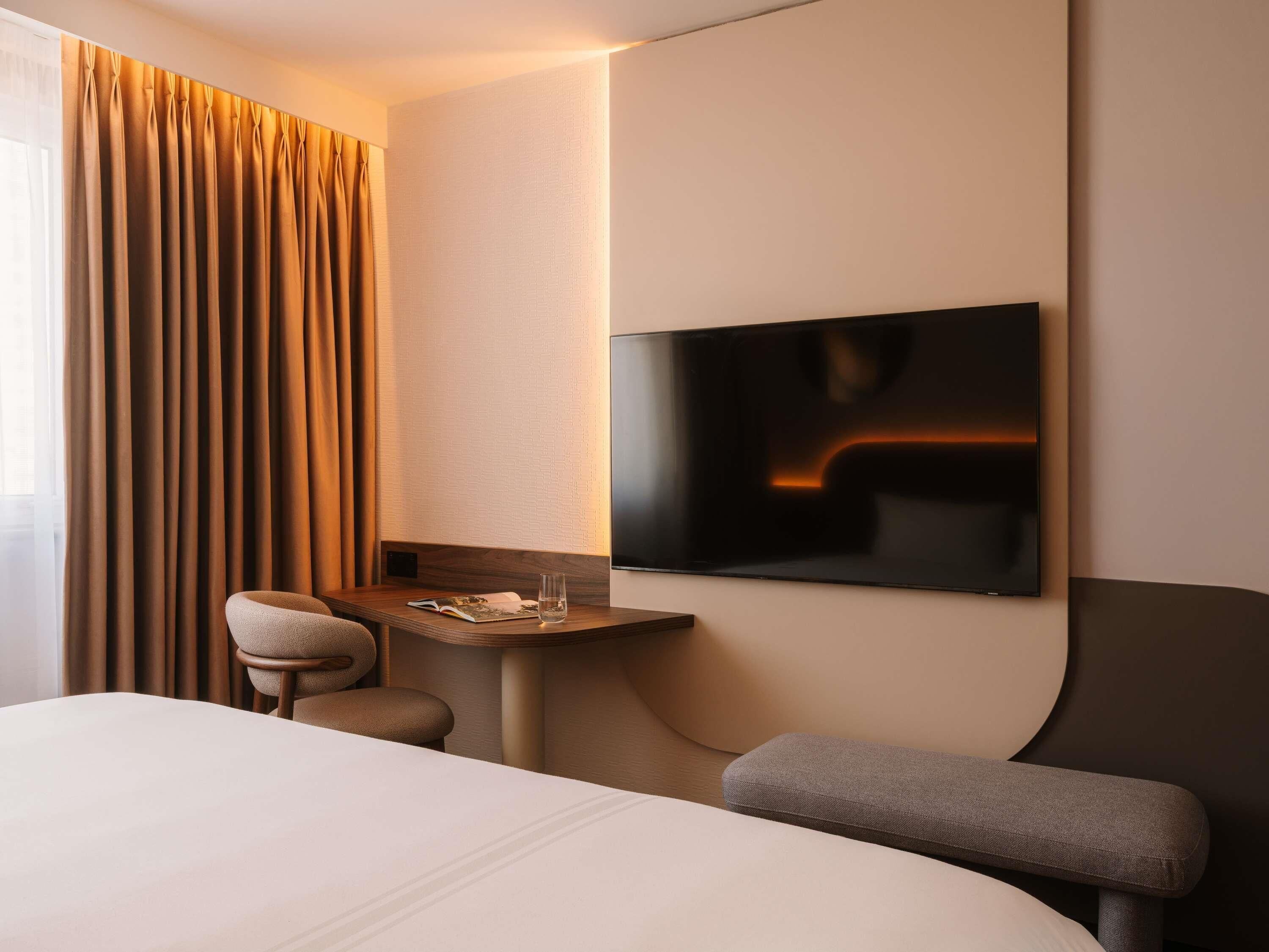 A hotel room with a bed, a desk, a chair, a TV, and a large window with curtains.