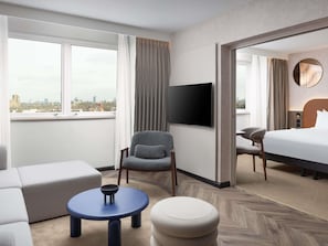 A hotel room with a city view, a flat-screen TV, a bed, a sofa, a chair, and a small round table. - Mercure London Earls Court (London)