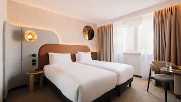 Ein Hotelzimmer mit zwei Betten, einem runden Spiegel, einem Nachttisch mit Lampe, einem Sessel und einem Fenster mit Vorhängen.