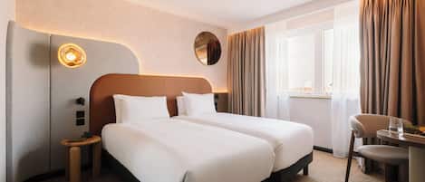 A hotel room with two beds, a round mirror, a bedside table with a lamp, a chair, and a window with curtains.