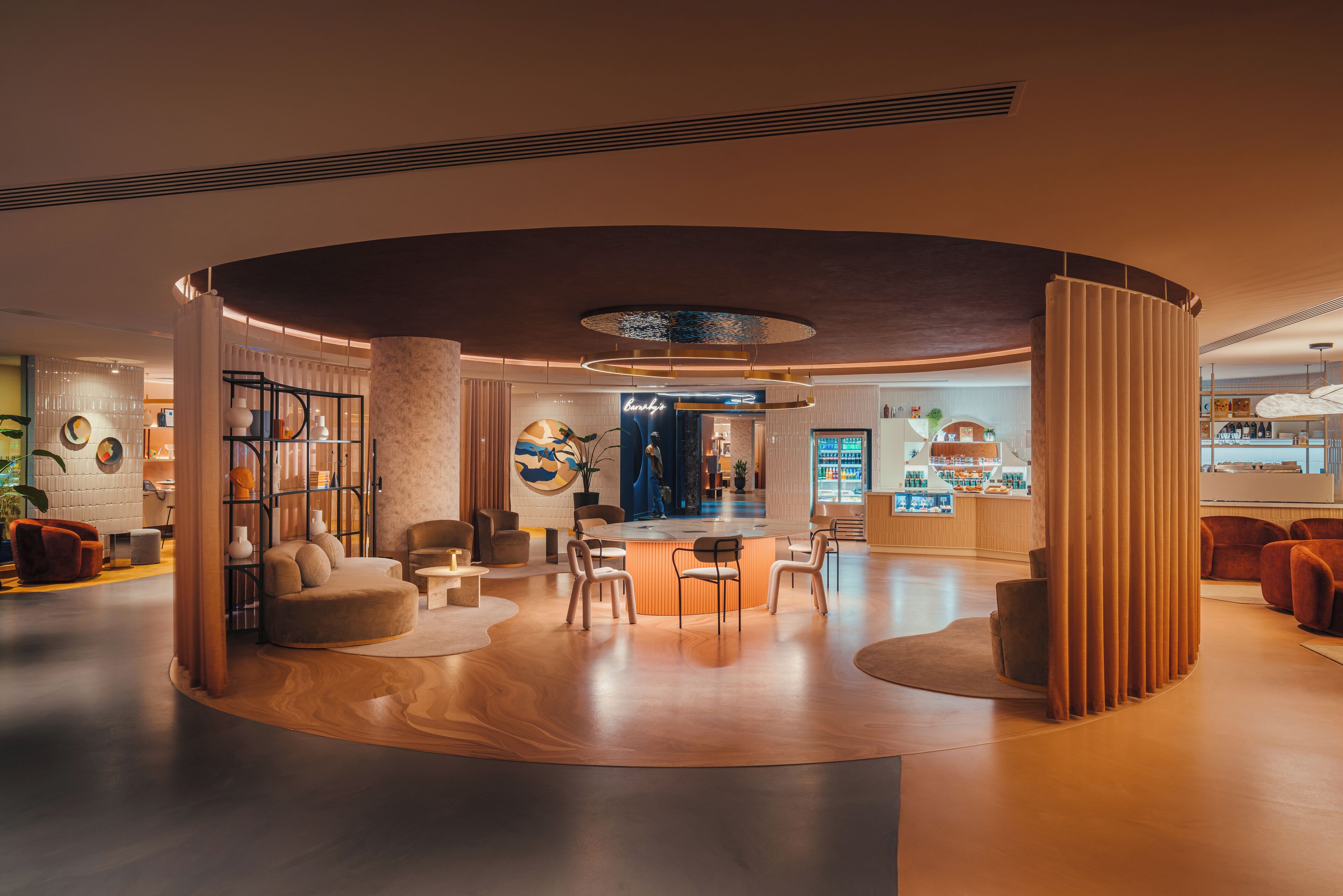 A modern hotel lobby with a central seating area, a reception desk, and a visible bar area.