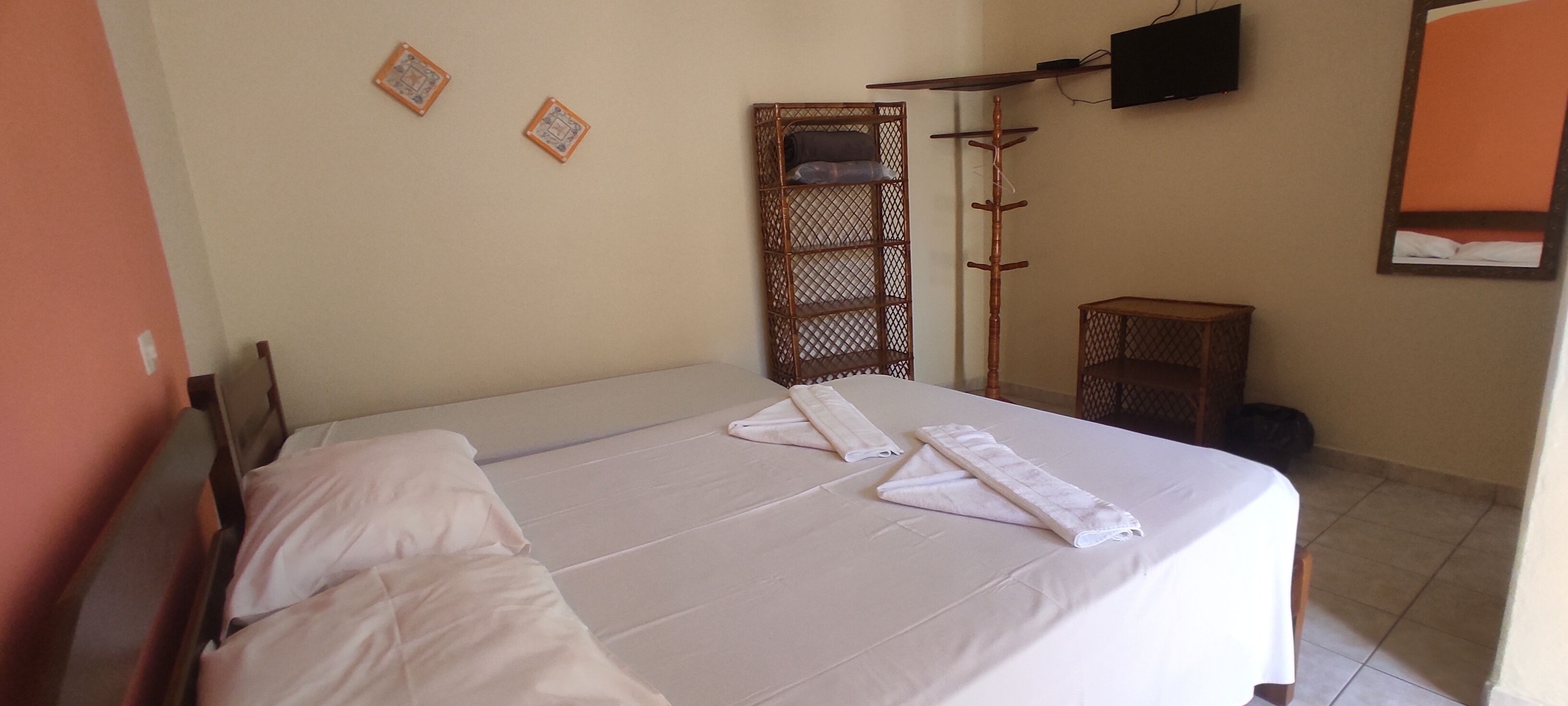 A neatly made bed with white linens and towels, a wooden wardrobe, a television mounted on the wall, and a small table with a bag on it.