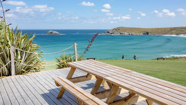 A wooden deck with a picnic table overlooking a coastal view with a grassy area and a rocky island in the distance. - A large three bedroomed apartment in West Pentire, Crantock. (Crantock)