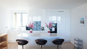 A modern kitchen with white cabinetry, a central island with bar stools, and a window providing natural light. - A large three bedroomed apartment in West Pentire, Crantock. (Crantock)