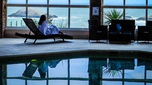 A person relaxing on a lounge chair by a large window overlooking a pool and ocean view. - A large three bedroomed apartment in West Pentire, Crantock. (Crantock)