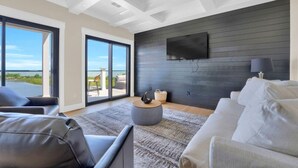 A modern living room with a large TV, comfortable seating, and a view through glass doors.