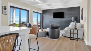 A modern living room with a large window, a flat-screen TV mounted on the wall, and a dining area with a white table and wicker chairs.