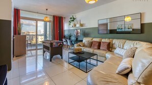 A modern living room with a sofa, coffee table, and a foosball table. There is a large window with red curtains, a view of the city, and a ceiling light fixture. - Designer, fully equipped 3BR home in Gzira w/WIFI (Il-Gżira)