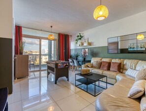 A modern living room with a sofa, coffee table, and a foosball table. There is a large window with red curtains, a view of the city, and a ceiling light fixture. - Designer, fully equipped 3BR home in Gzira w/WIFI (Il-Gżira)