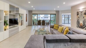 A modern living room with a grey sectional sofa, a flat-screen TV, a glass coffee table, and a dining area with chairs. - Designer, luxurious 3BR home in Pender Gardens (San Ġiljan)