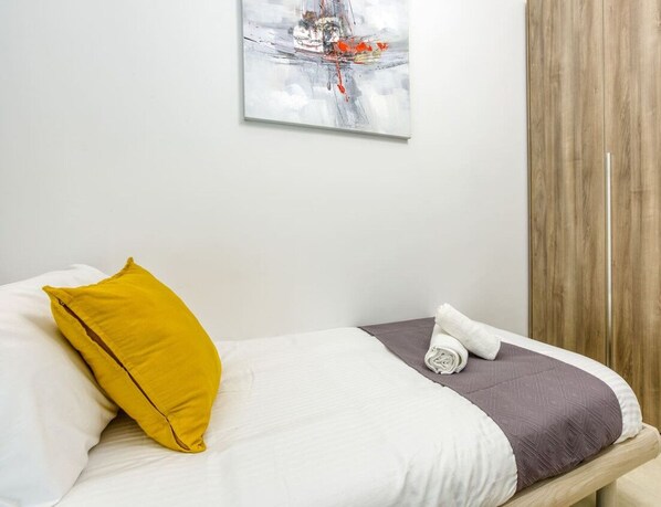 A neatly made bed with a white comforter, a yellow pillow, and a rolled white towel.