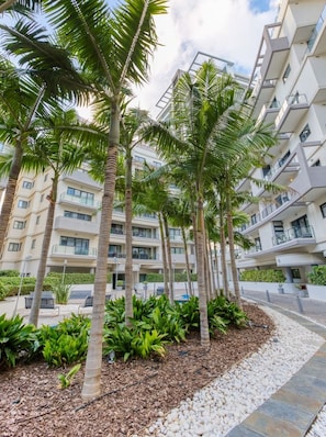 Um edifício residencial moderno, de vários andares, com varandas, rodeado por palmeiras e um jardim bem cuidado.