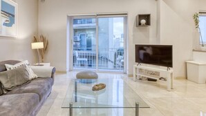 Ein modernes Wohnzimmer mit einem Glastisch, einem grauen Sofa, einem Flachbildfernseher auf einem weißen Schrank und einem Balkon mit Blick auf die Stadt.