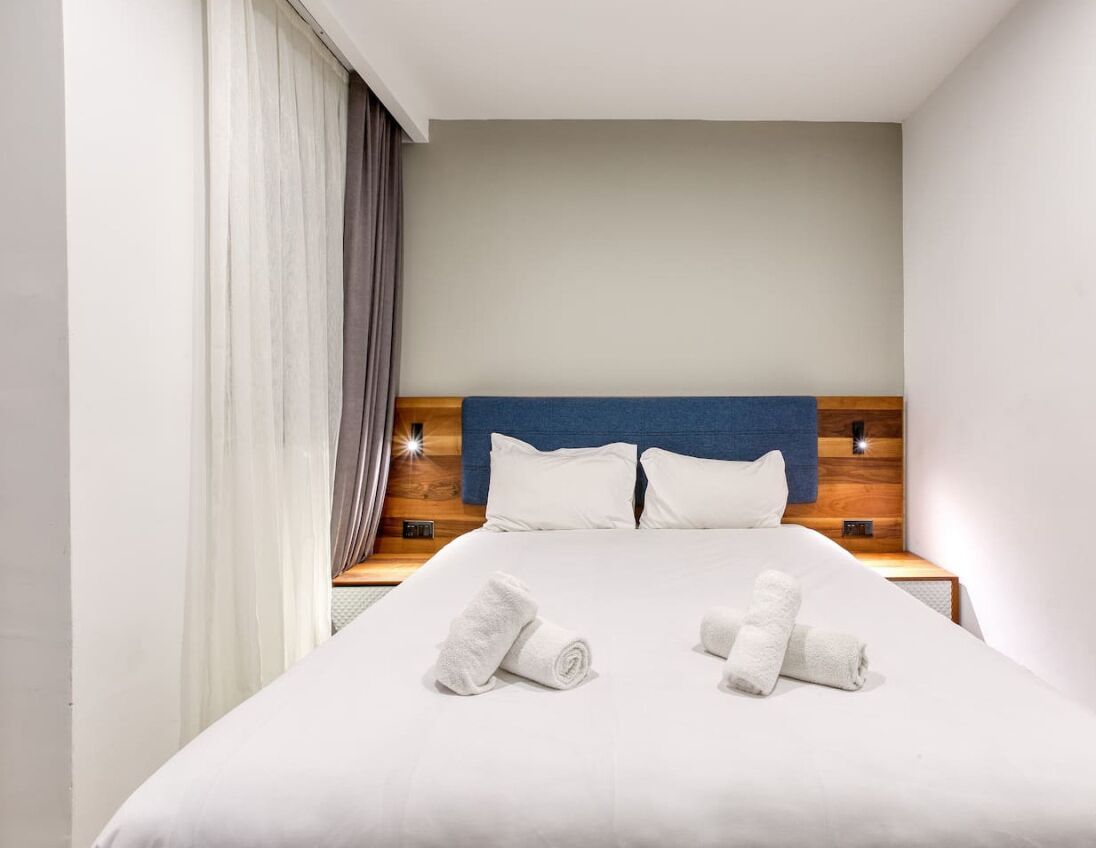 Una cama bien tendida con sábanas blancas y dos toallas enrolladas, un cabecero de madera y una mesita de noche con una lámpara.
