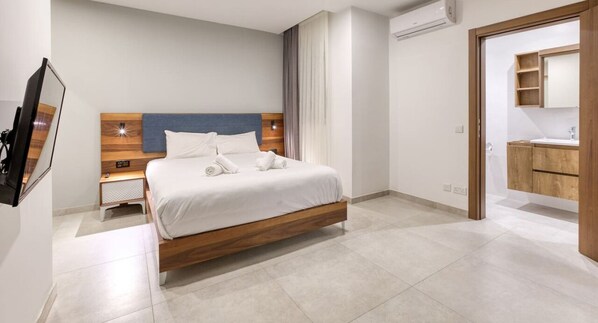 Una habitación de hotel moderna con una cama de madera, un televisor de pantalla plana, una mesita de noche y un baño con tocador y espejo de madera.