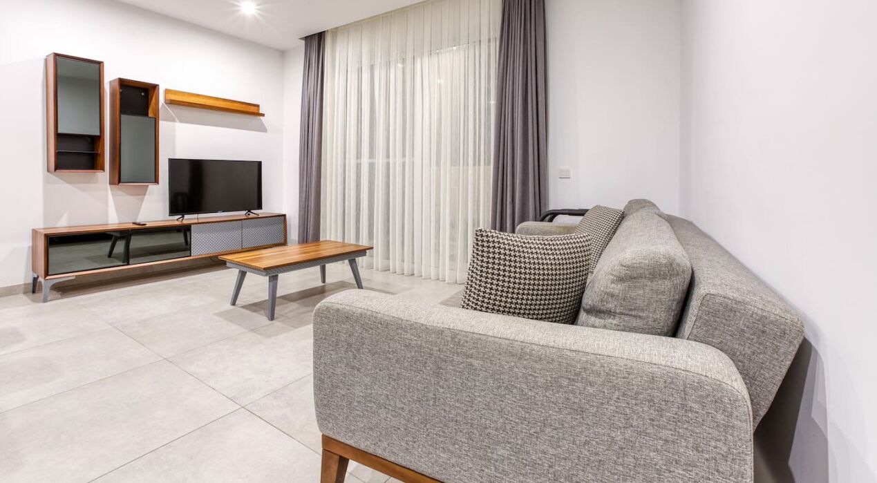 Una sala de estar moderna con un sofá gris, una mesa de centro de madera, un televisor sobre un mueble de madera y un ventanal con cortinas translúcidas.