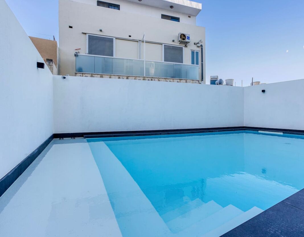 Une piscine sur le toit, avec une eau d’un bleu cristallin, entourée de murs blancs et d’un bâtiment moderne doté de grandes fenêtres et d’un balcon.