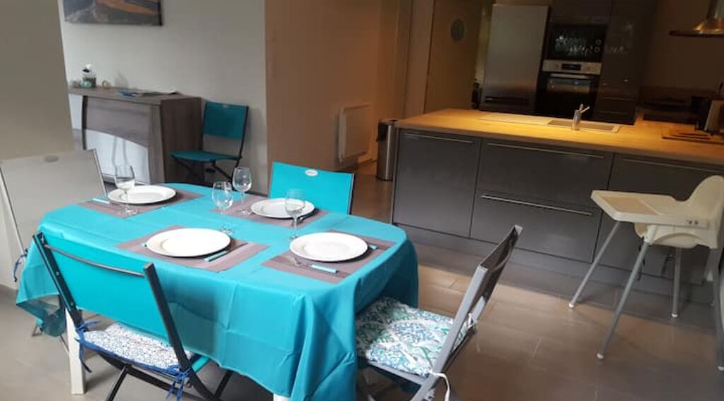 Un coin repas avec une table dressée pour quatre personnes, une cuisine en arrière-plan et une chaise blanche sur la droite.