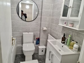 A modern bathroom with grey tiles, a white toilet, a small trash bin, a white sink with toiletries, and a mirror. - London - Sleeps 4 - Parking - Garden - Petfriendly (London)