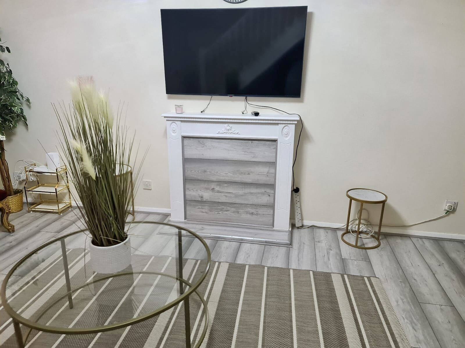 A living room with a fireplace, a flat-screen TV mounted above it, a round glass coffee table, and a striped rug on the floor.