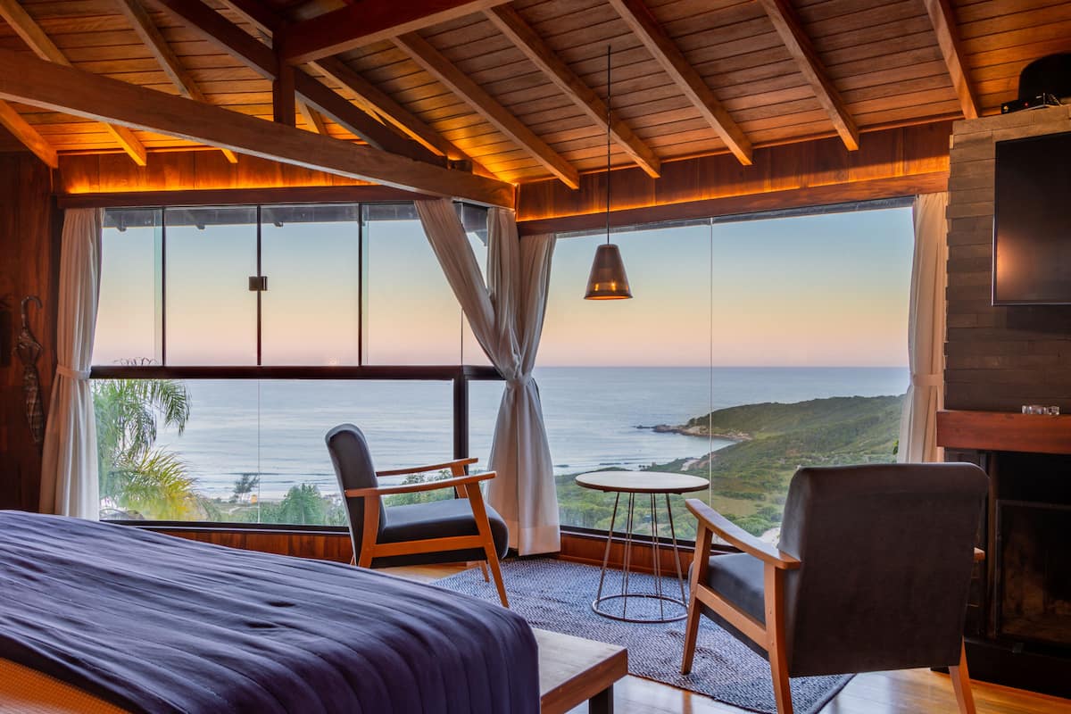 A room with a bed, a TV, a fireplace, a view of the ocean, and a table with chairs.