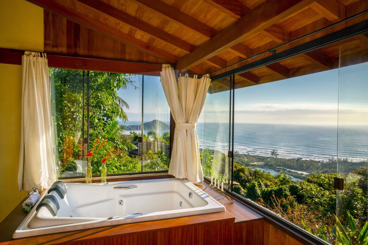 A modern bathroom with a large bathtub, wooden ceiling, and a view of the ocean and greenery.